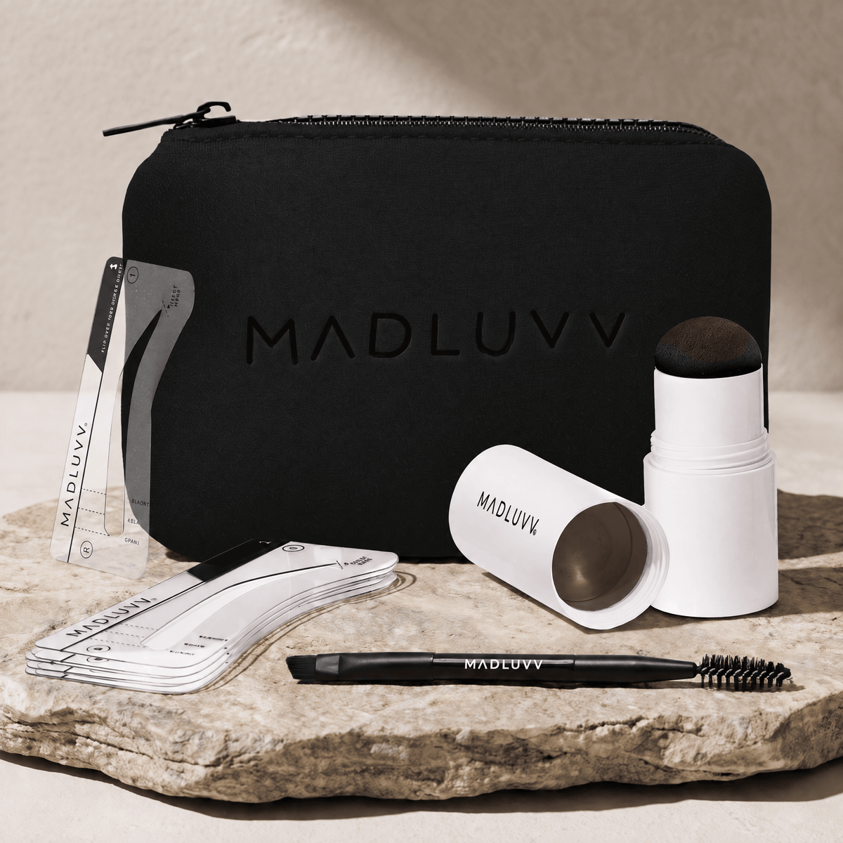A Madluvv brow kit arranged on a stone slab against a neutral beige background. The set includes a black zippered pouch with embossed branding, a brow stamp, a white and black spoolie applicator, clear and white stencil templates sheets