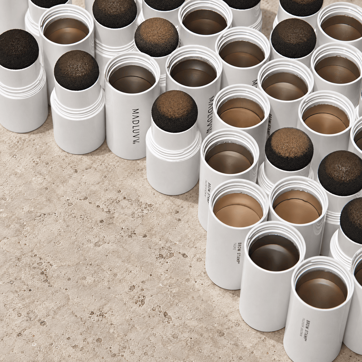Multiple open white cylindrical makeup pots arranged on a light beige stone surface, showing pomeade products in shades of brown, taupe, and dark brown. The brand name 'Madluvv' is visible on some containers.