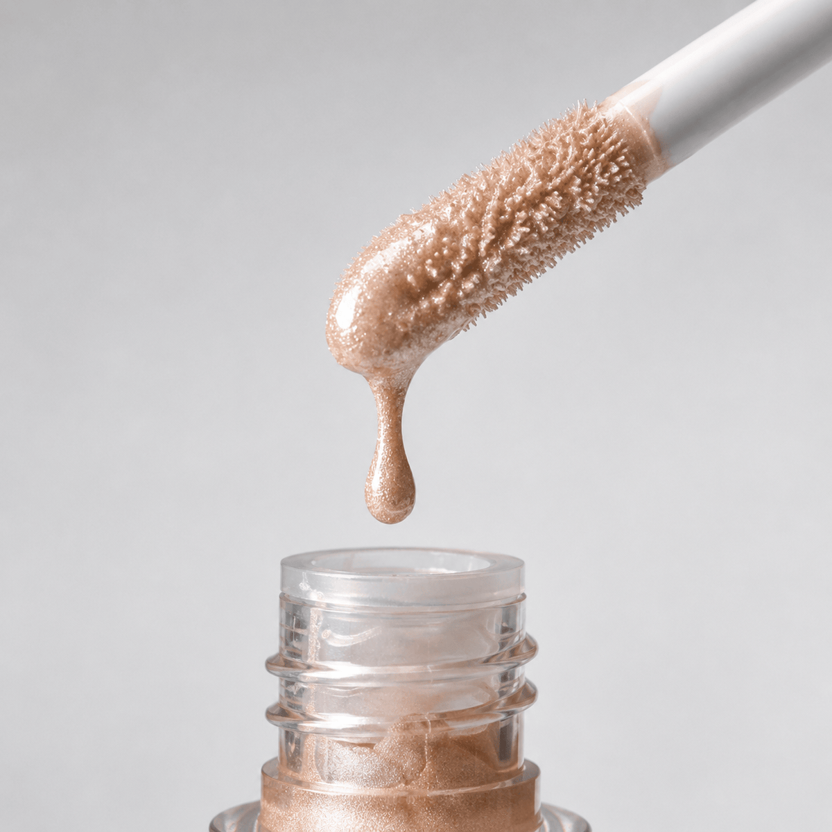 A close-up of a glittery nude-beige liquid cosmetic, shown dripping from a doe-foot applicator onto an open clear bottle against a light gray background.