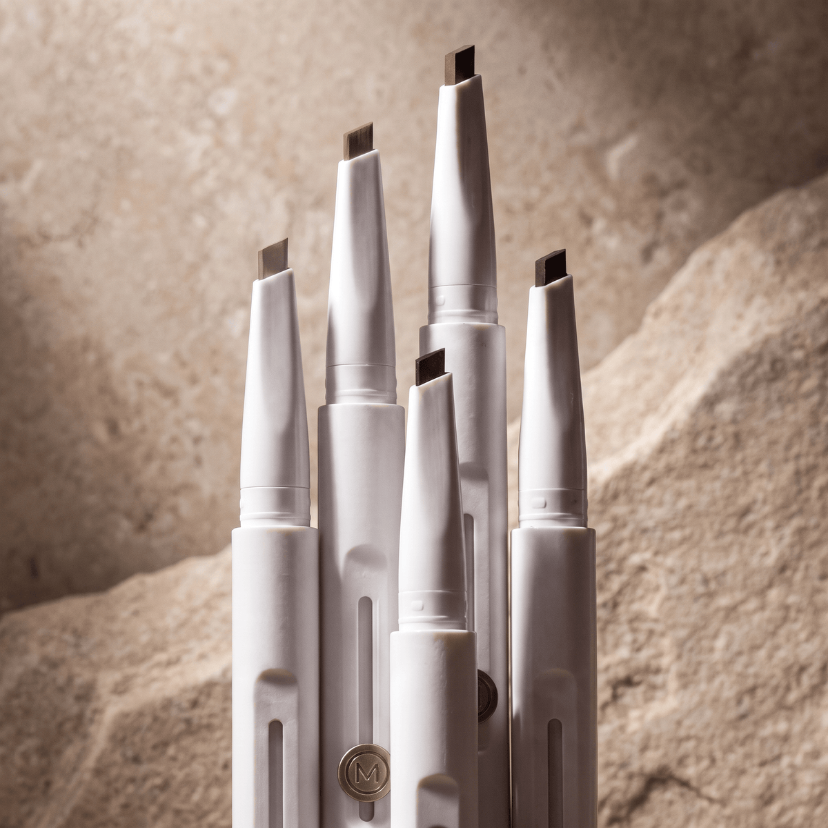Five white cosmetic pencils with dual-ended tips arranged together against a textured beige stone background.