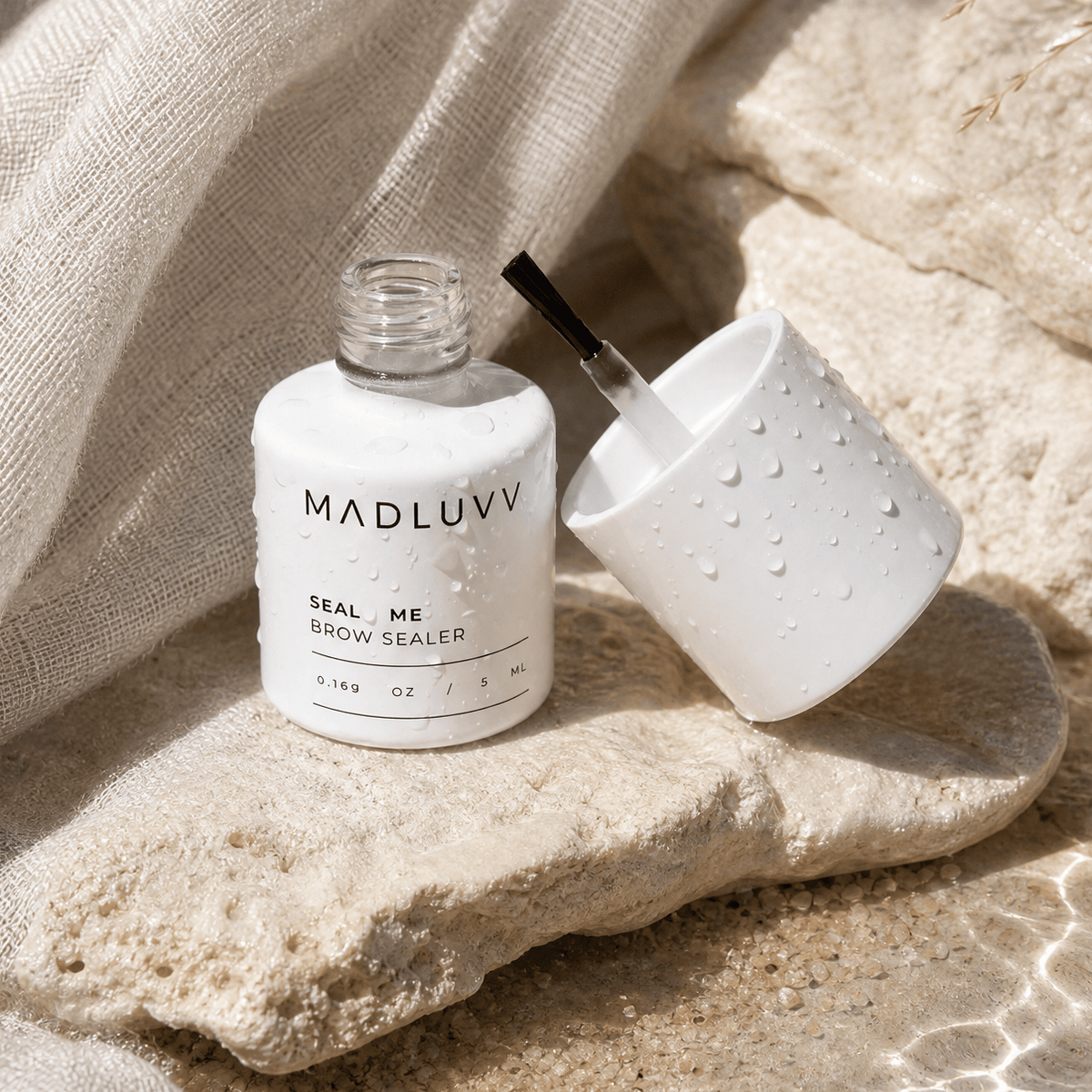Bottle of MADLUUV eyebrow sealer and applicator on a stone surface with fabric in the background