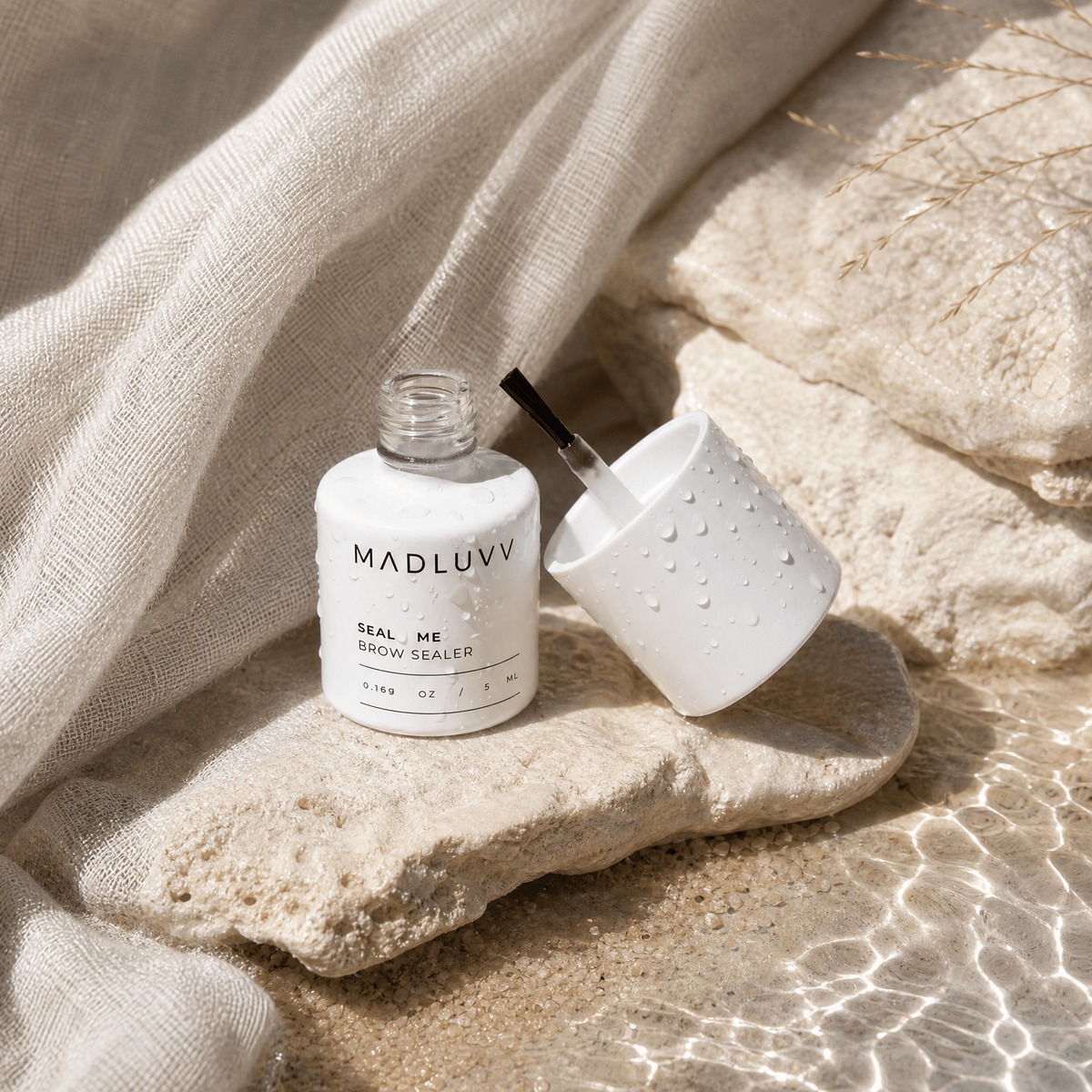 Bottle of MADLUUV eyebrow sealer and applicator on a stone surface with fabric in the background